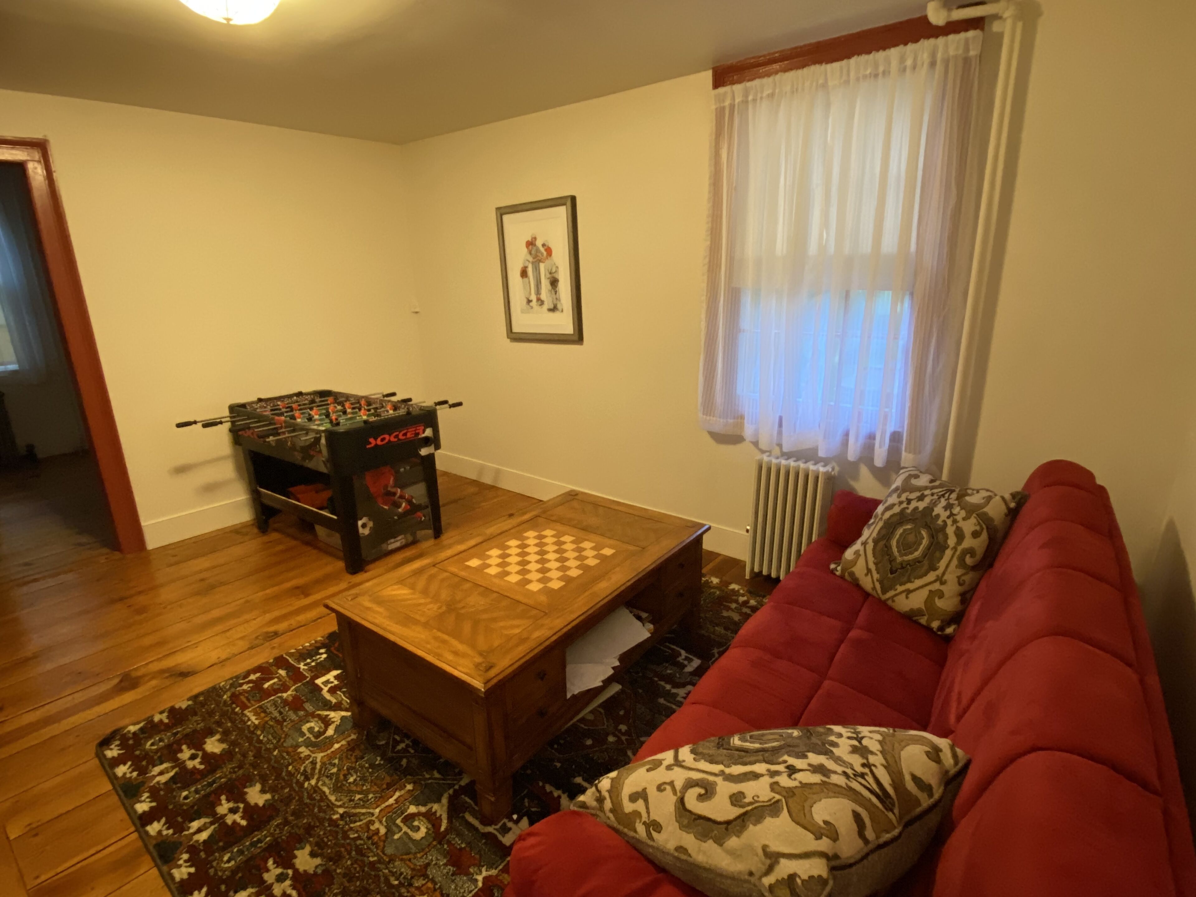 Una sala de estar con un sofá rojo, una mesa de centro de madera, una mesa de fútbol de mesa, un cuadro en la pared y una alfombra estampada.