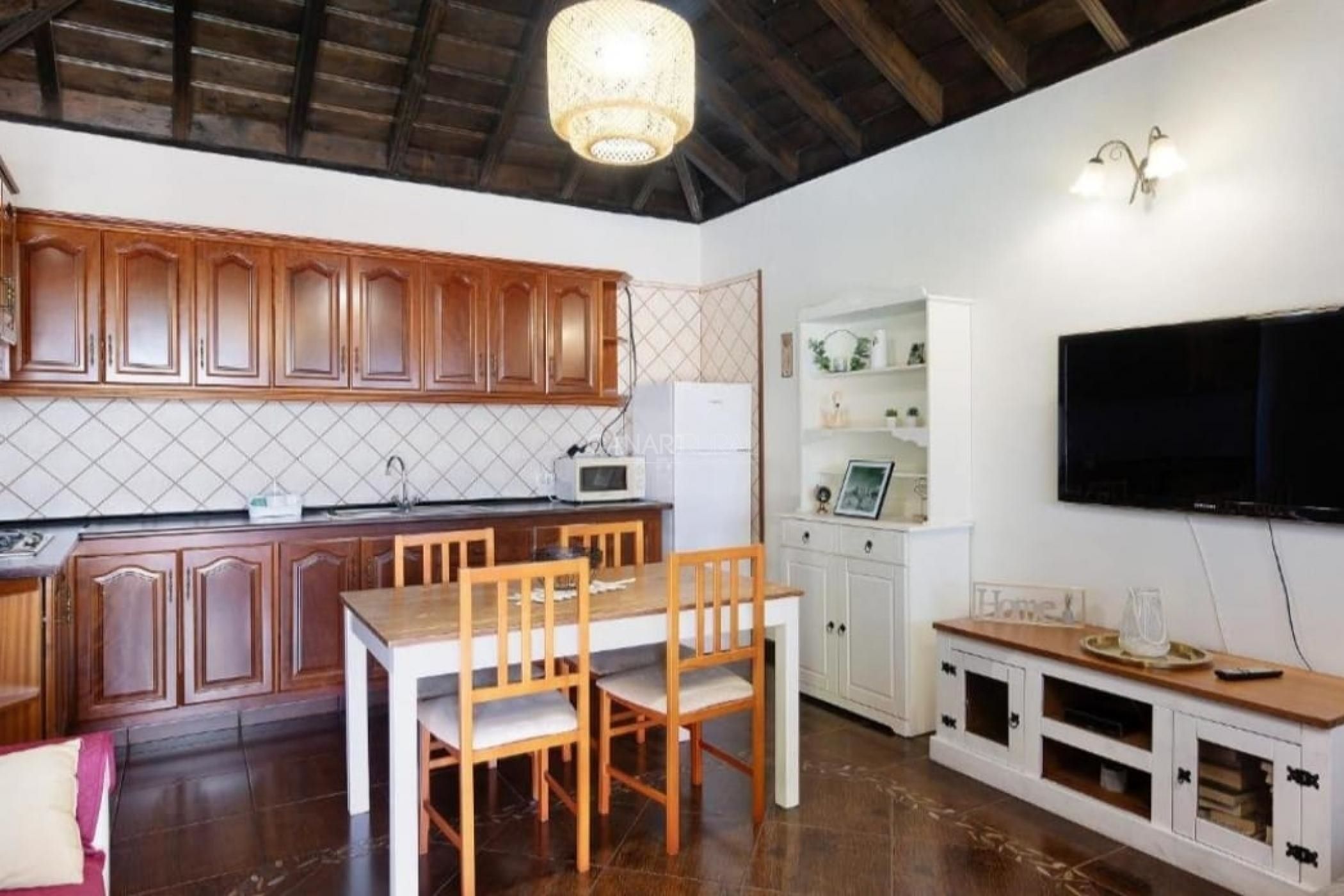 Une cuisine avec des meubles en bois, une table à manger, des chaises, un four à micro-ondes et un téléviseur fixé au mur.