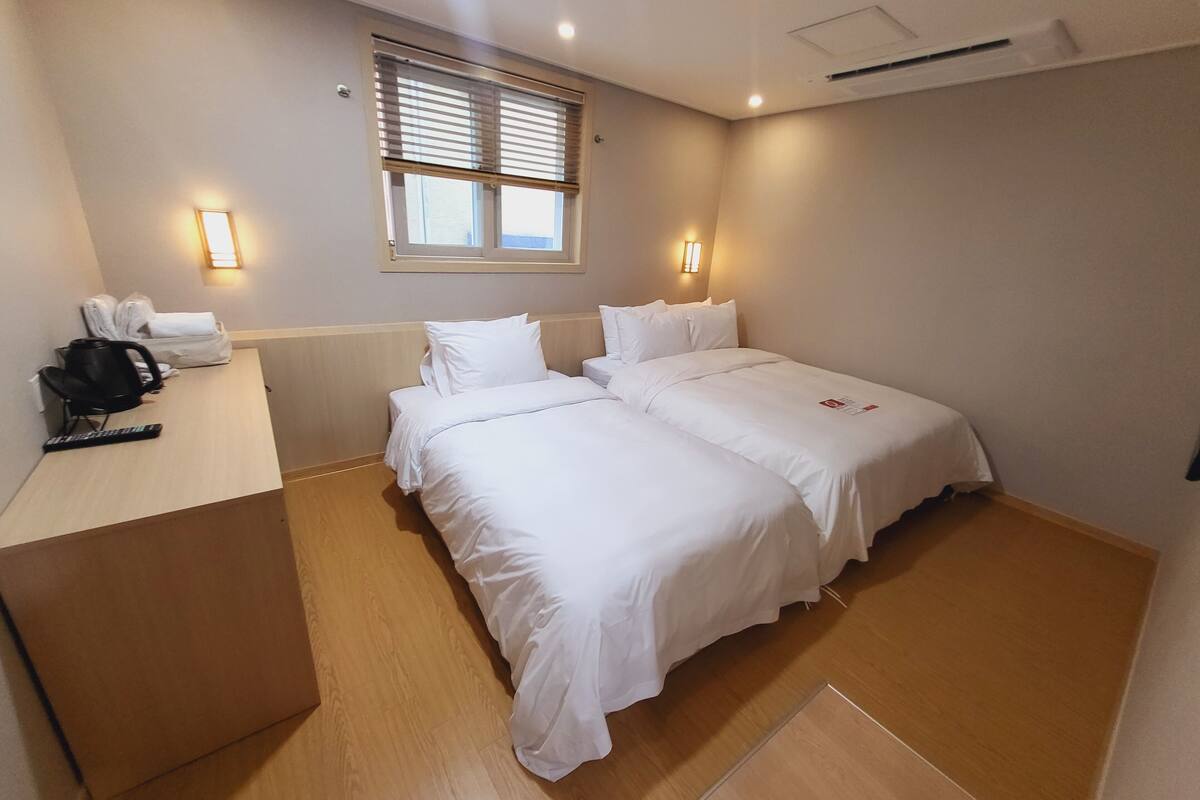 A hotel room with two beds, a desk with a chair, a telephone, and a window with blinds.