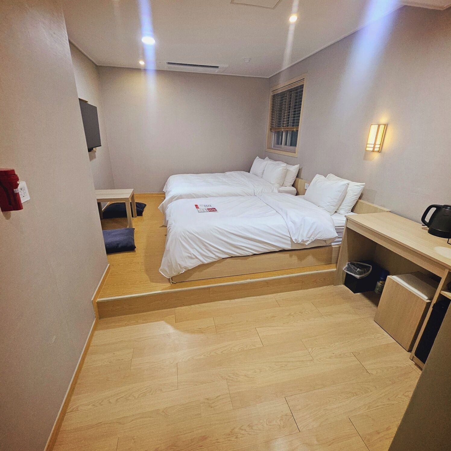 A hotel room with two single beds, a small desk, a chair, a trash bin, and a window with blinds.