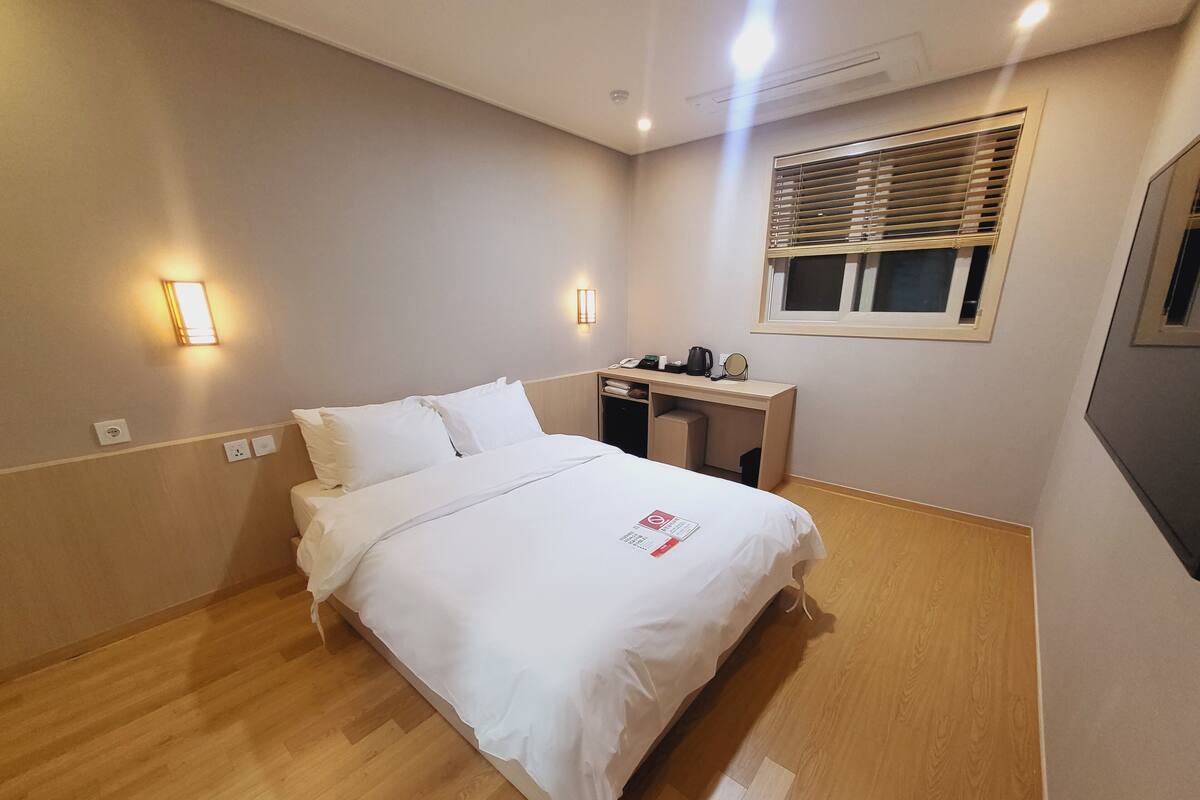 A hotel room with a bed, bedside table, lamp, and a window with blinds.