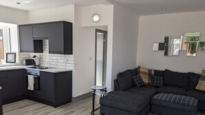 A modern kitchen with dark cabinetry and a built-in oven, a gray sectional sofa with plaid cushions, and a small round table. - Lovely 3-bed House in Lytham Saint Annes (Lytham St. Anne's)