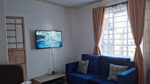 A living room with a blue sofa, a TV mounted on the wall, and a window with curtains. - Cozy studio with WIFI &Parking in South B (Nairobi)