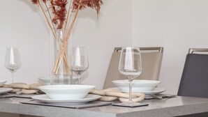 Een eettafel gedekt met witte kommen, glanzende wijnglazen en een vaas met gedroogde planten.