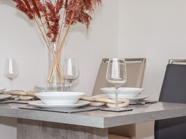 Una mesa de comedor dispuesta con cuencos blancos, copas de vino transparentes y un florero con plantas secas.