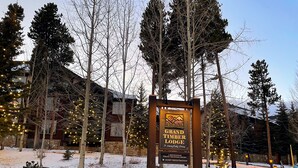 Placa para Grand Timber Lodge com base de pedra, rodeada por terreno e árvores cobertas de neve.