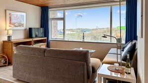 A living room with a sofa, a TV, a small table with a bottle, and a view of the beach. - Lovely apartment in Bergen aan Zee (Bergen aan Zee)