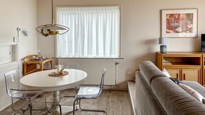 A cozy living room with a round dining table, clear chairs, a sofa, a wooden cabinet, and a window with sheer curtains. - Lovely apartment in Bergen aan Zee (Bergen aan Zee)
