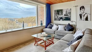 A modern living room with a glass coffee table, grey sofas, and a view of the beach. - Lovely apartment in Bergen aan Zee (Bergen aan Zee)