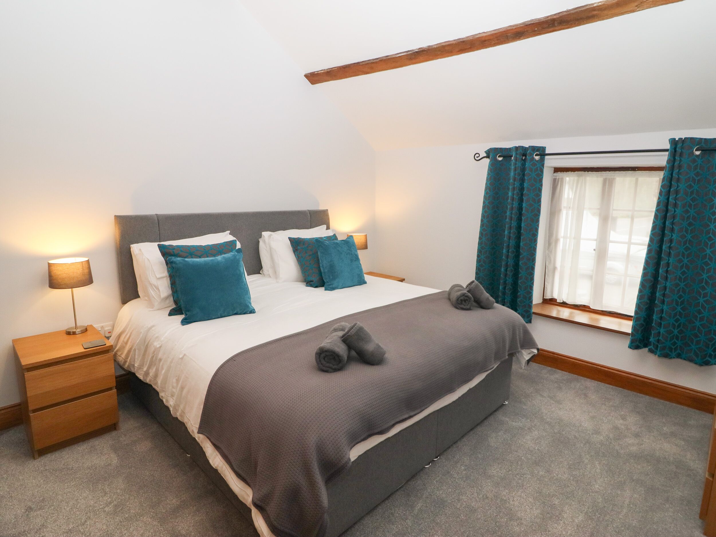 A bedroom with a bed, nightstands, a lamp, a window with curtains, and a wooden beam above the bed.