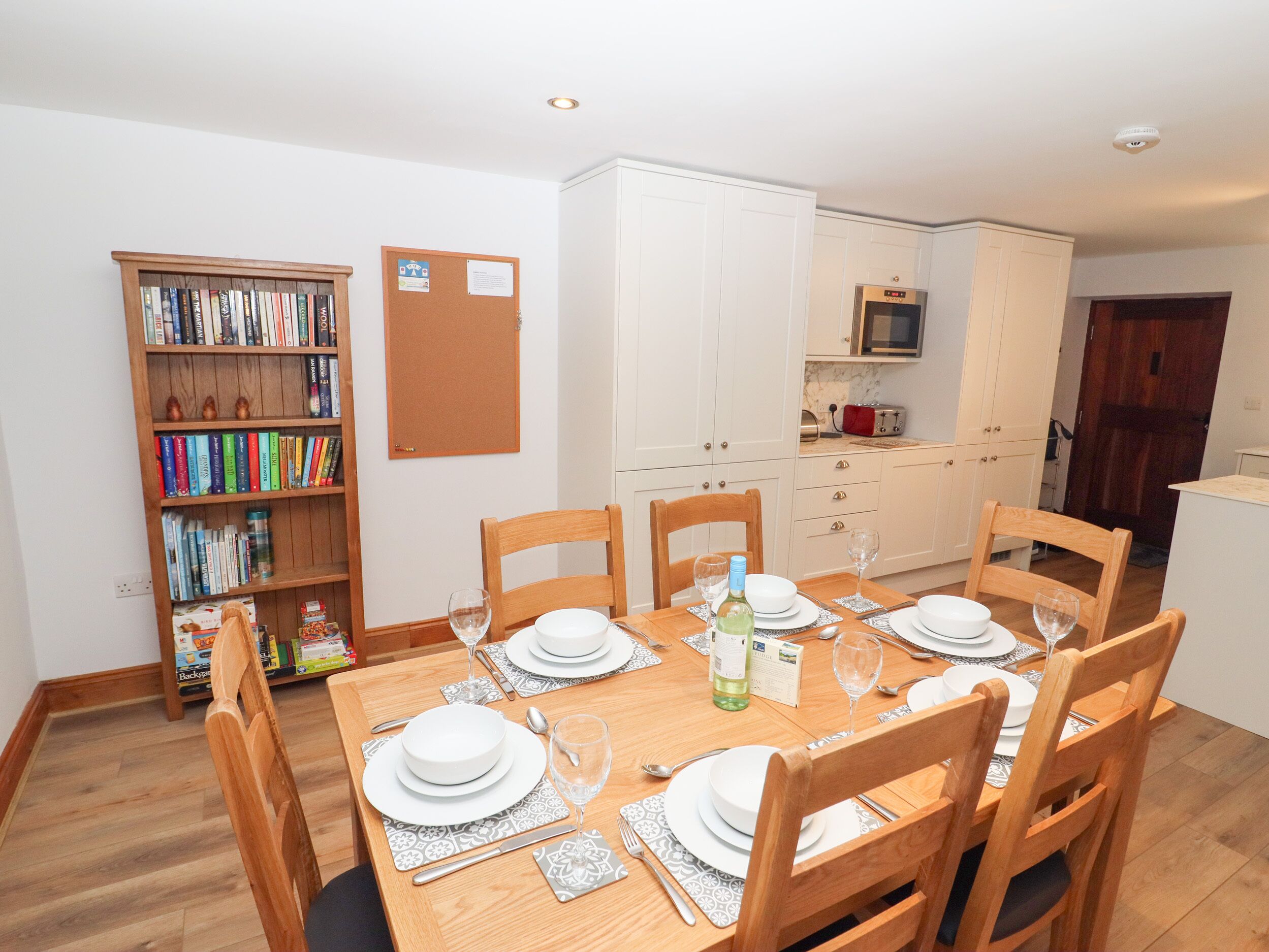 Comedor con una mesa de madera dispuesta para cuatro personas, rodeada de sillas. A la izquierda, una estantería llena de libros.