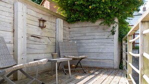 Une terrasse en bois avec deux fauteuils en osier et une petite table, entourée d’une clôture en bois et de verdure.