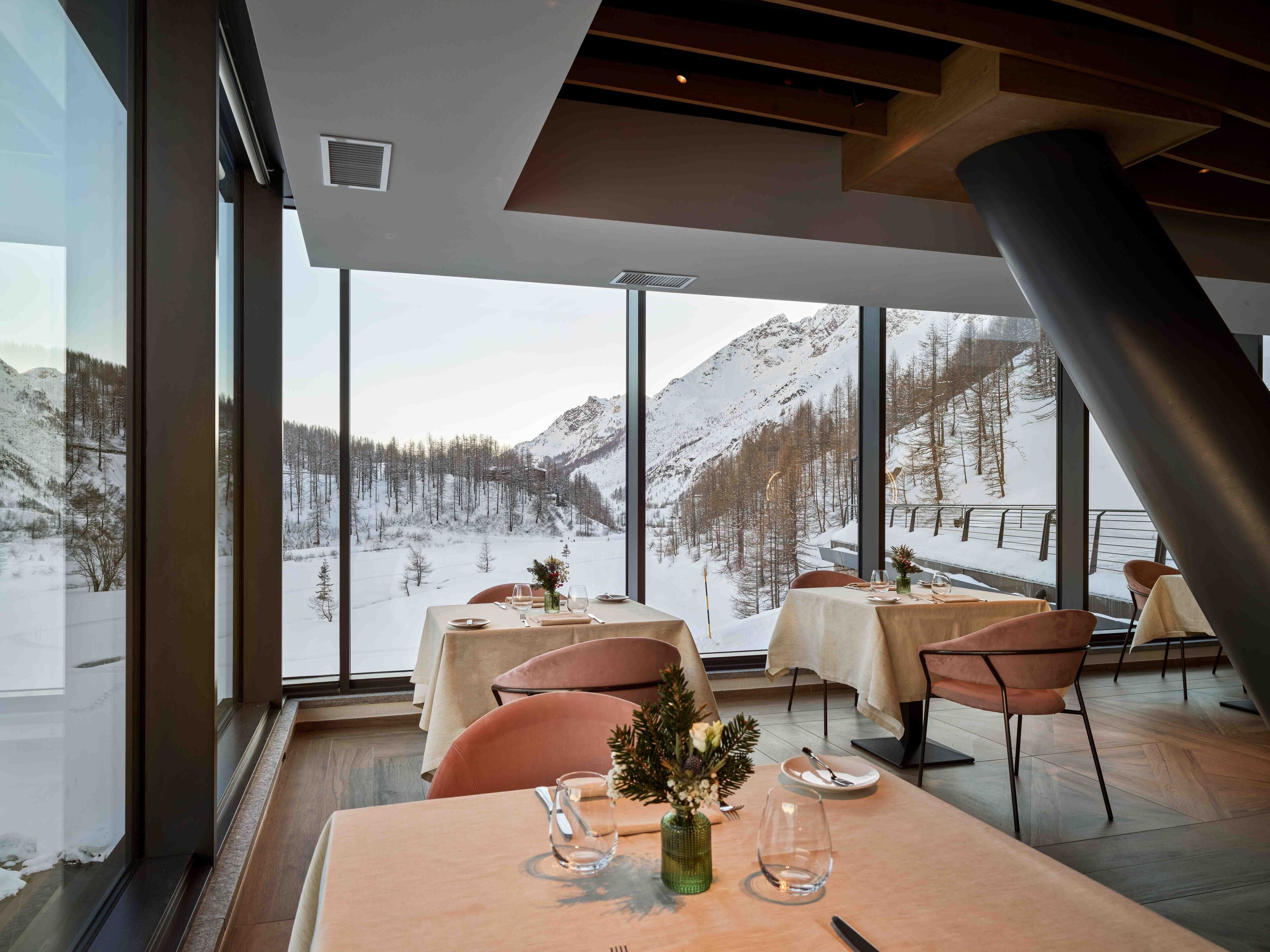 A dining area with tables set for two, large windows offering a view of a snowy mountain landscape.