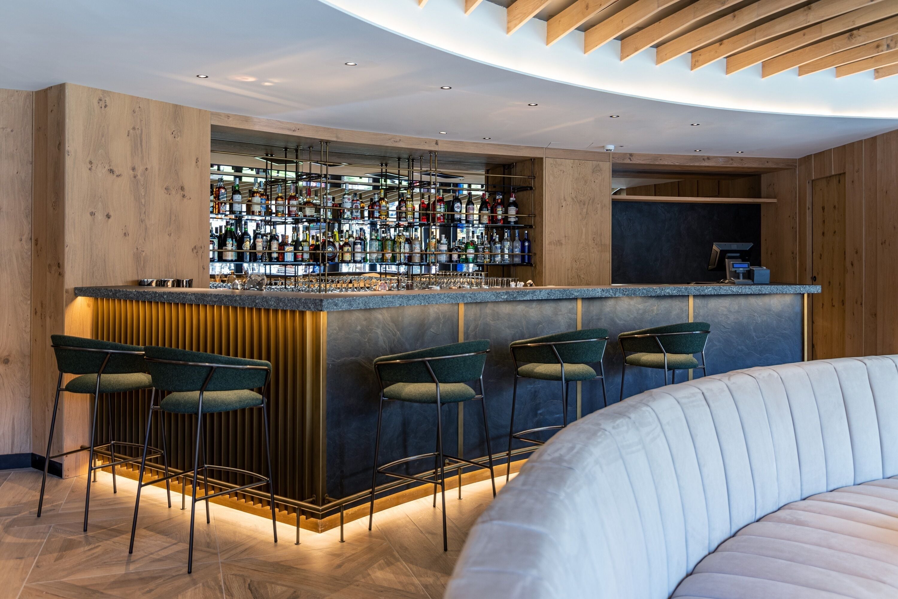 Un área de bar moderna con mostrador de madera, varios taburetes y una gran variedad de botellas.
