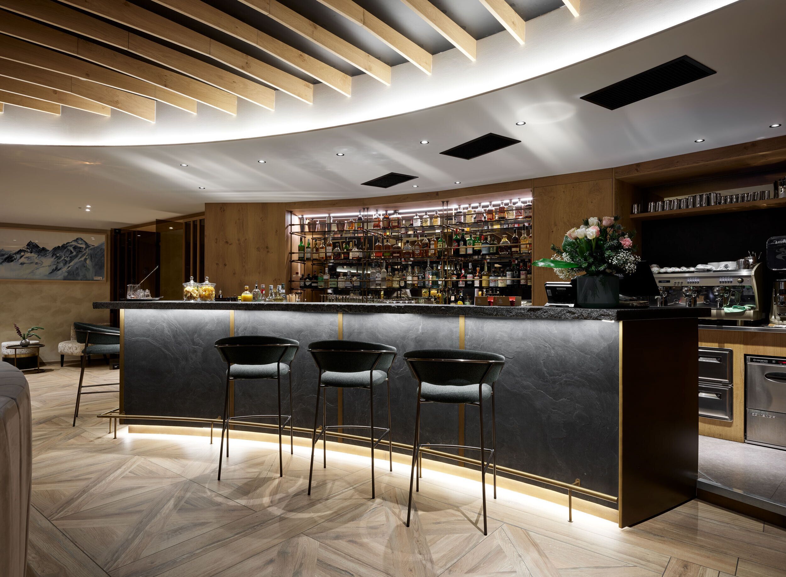 A modern bar area with a curved counter, three bar stools, and a well-stocked liquor shelf.