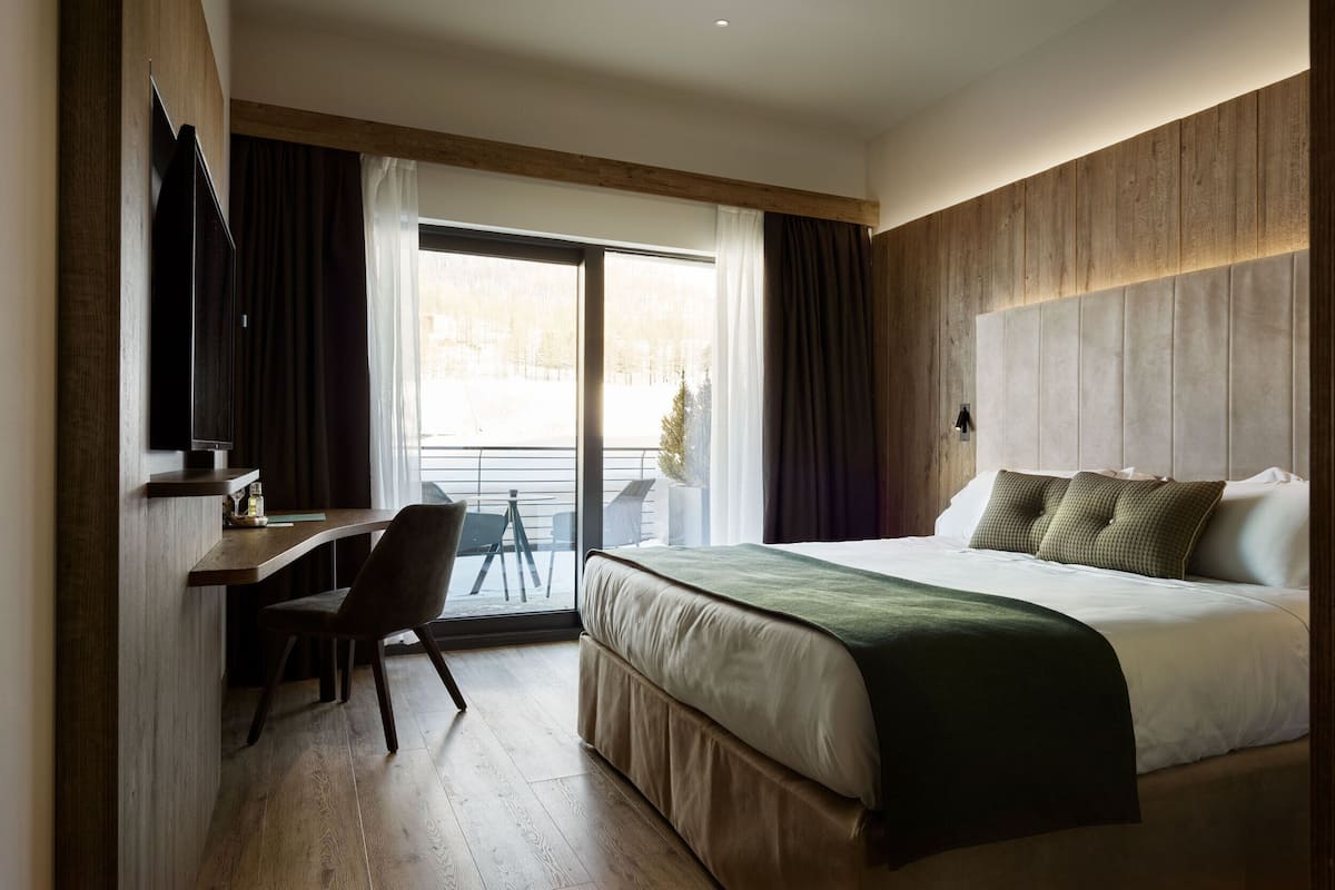 Quarto de hotel com cama, escrivaninha e cadeira. Há uma janela com cortinas e vista para árvores lá fora.