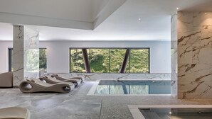 Een modern binnenzwembad met marmeren wanden en vloer, grote ramen met uitzicht op groen, en comfortabele ligbedden.