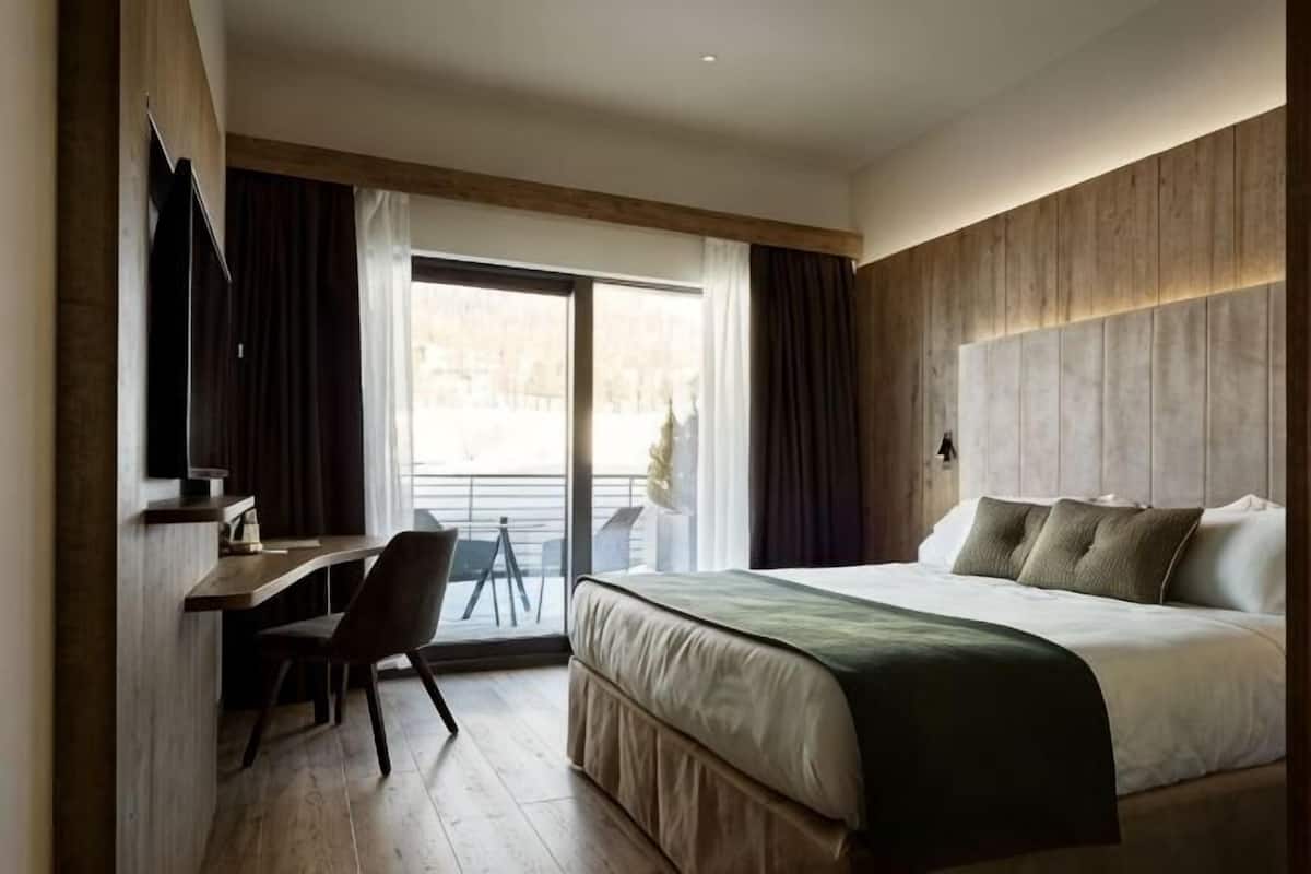Quarto de hotel com cama, escrivaninha, cadeira, televisão e varanda com cortinas.