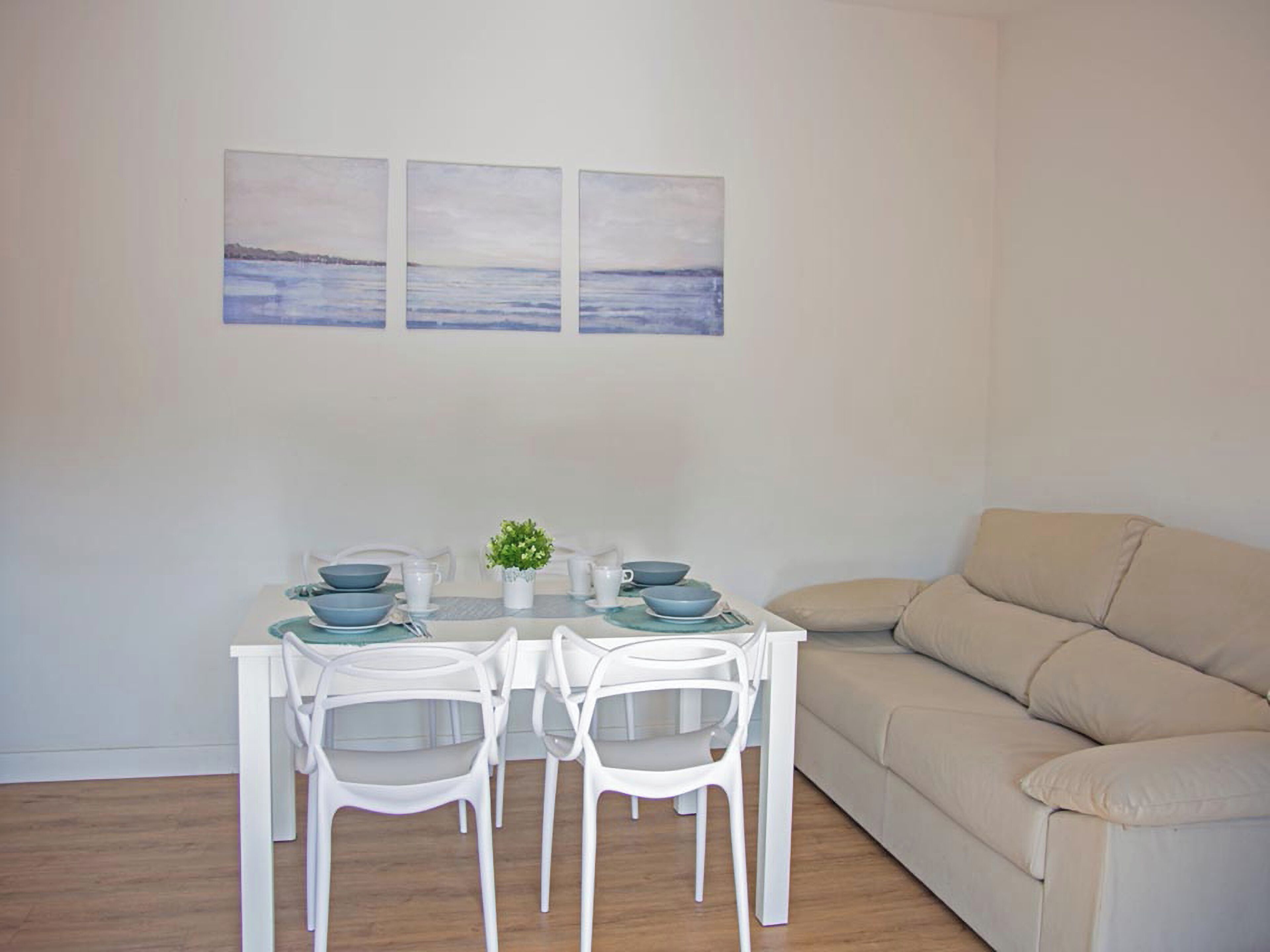 Comedor con una mesa dispuesta para cuatro personas, sillas blancas, un sofá beige y tres cuadros colgados en la pared.
