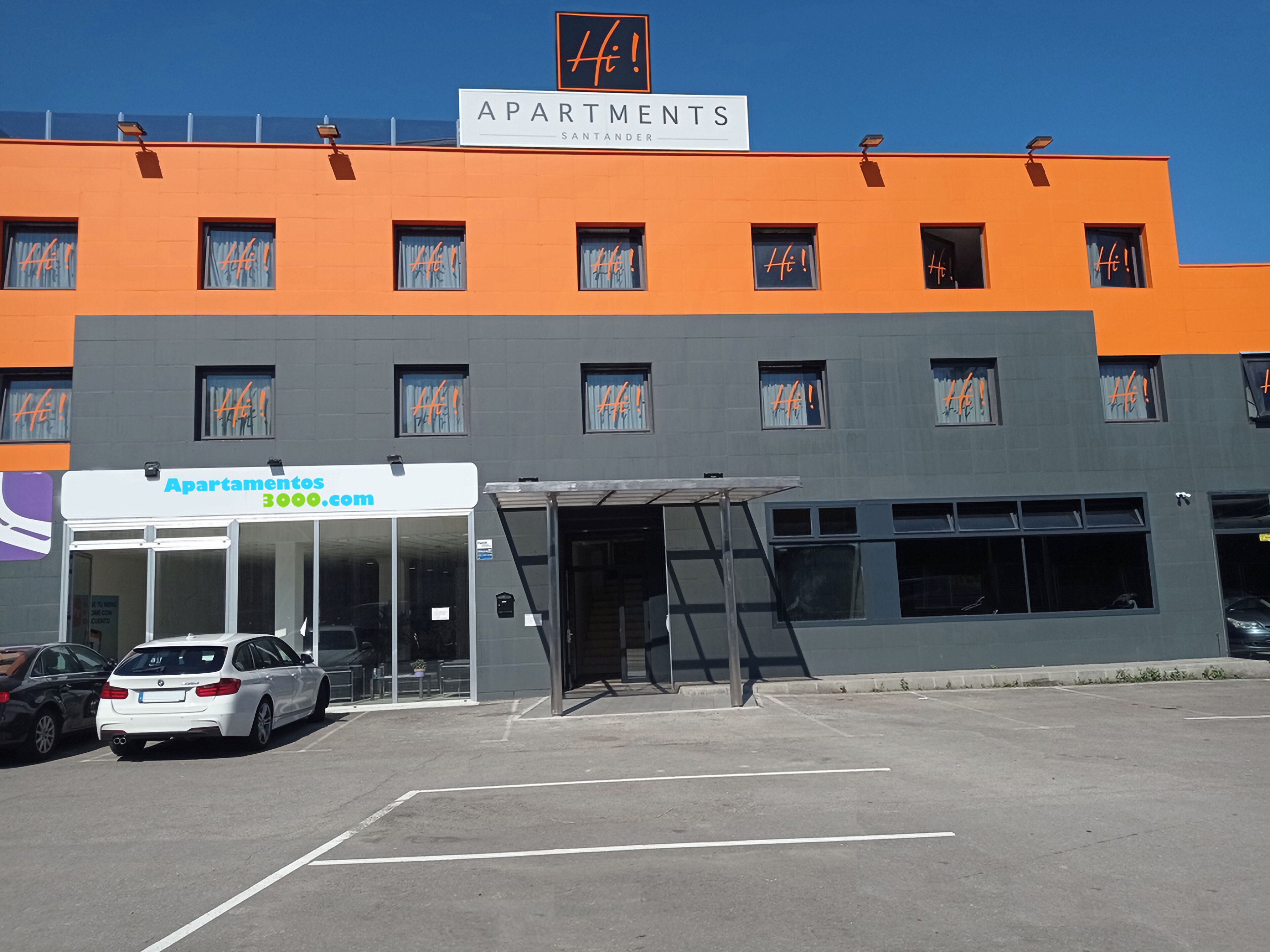 Edifici modern amb un rètol que diu 'Apartments Santander' i una direcció web 'Apartments3000.com'. Hi ha un cotxe blanc estacionat a la façana.
