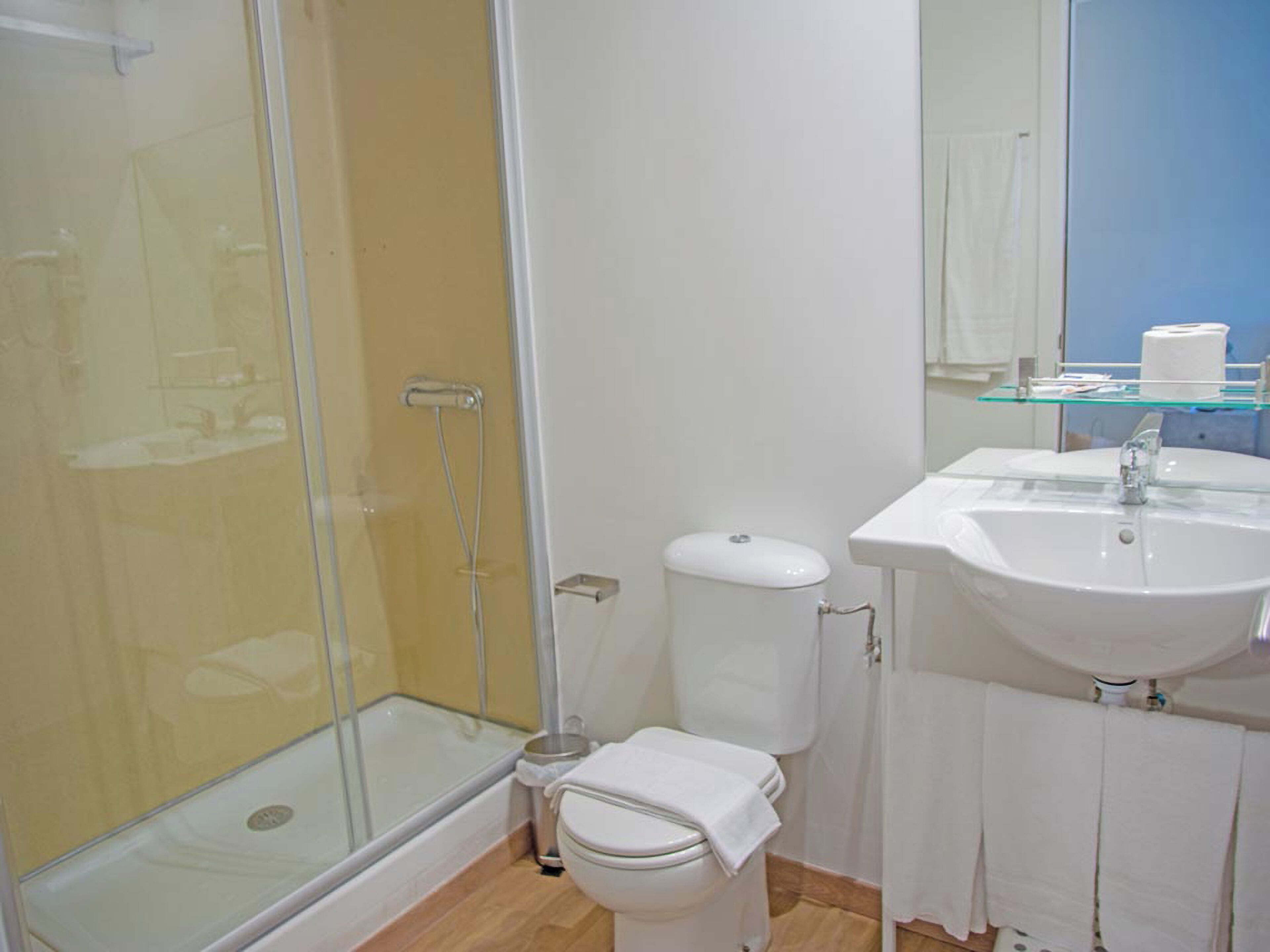 Un baño moderno con cabina de ducha de vidrio, inodoro blanco y lavamanos con repisa de vidrio arriba.