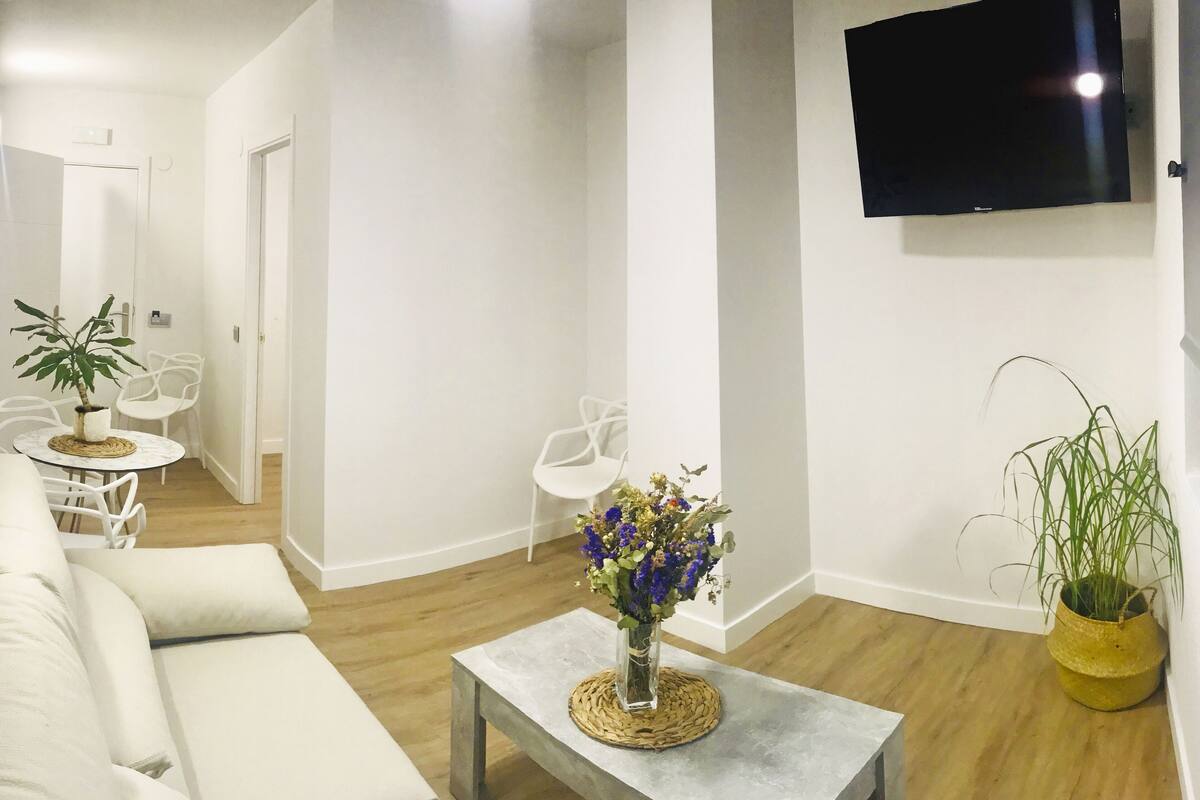 Una sala de estar moderna con un sofá, una mesa de centro con un florero, un televisor montado en la pared y una planta en maceta.