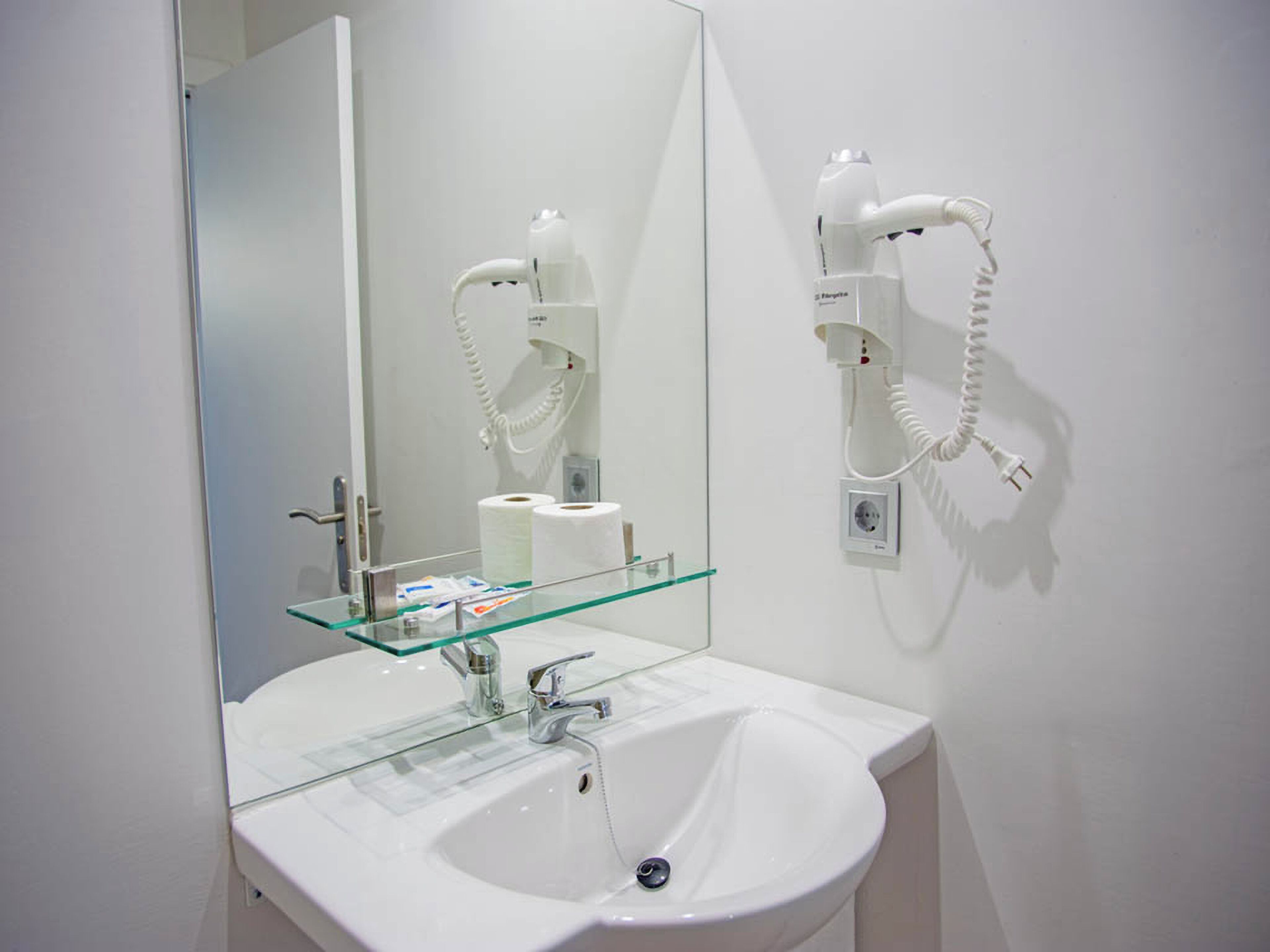 Une salle de bain avec un lavabo blanc, une étagère en verre, un miroir et un sèche-cheveux fixé au mur.