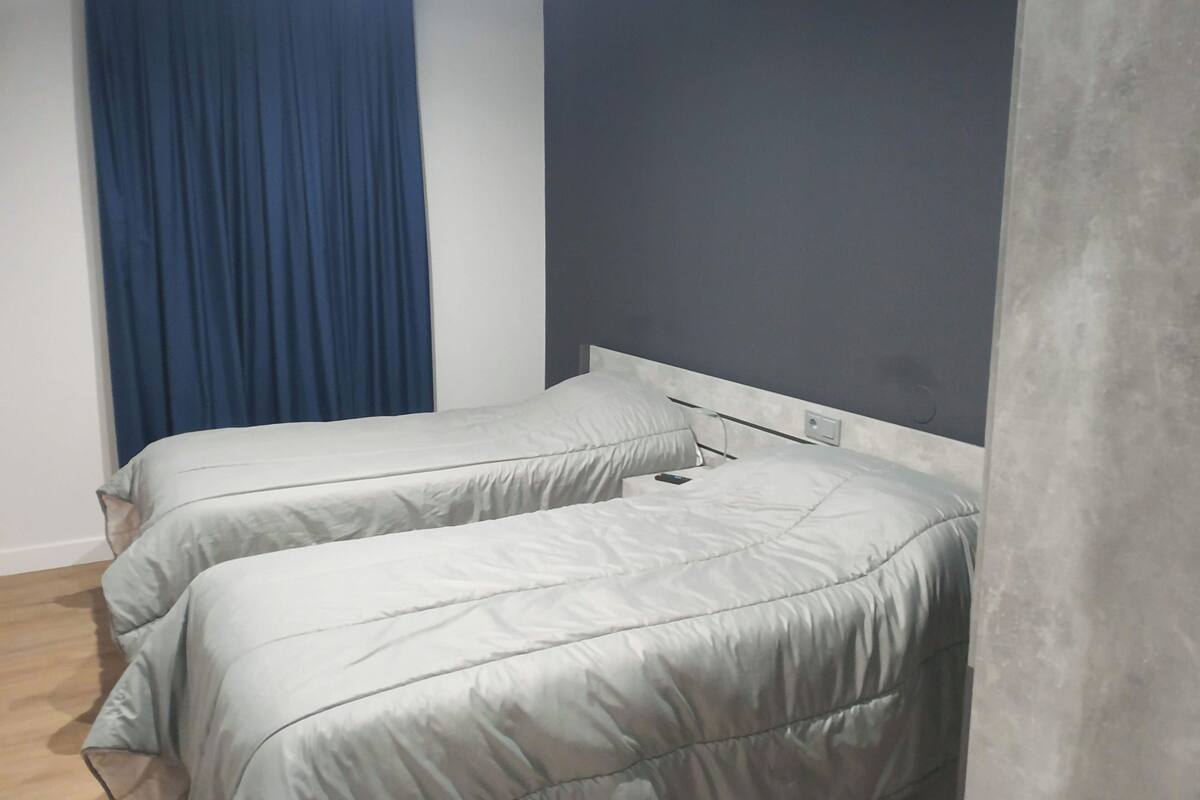 Un dormitorio pequeño y moderno con dos camas, una pared oscura y una cortina azul.