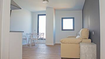 Une pièce avec un plancher en bois, un canapé, une petite table et deux chaises. Il y a une fenêtre et une porte.