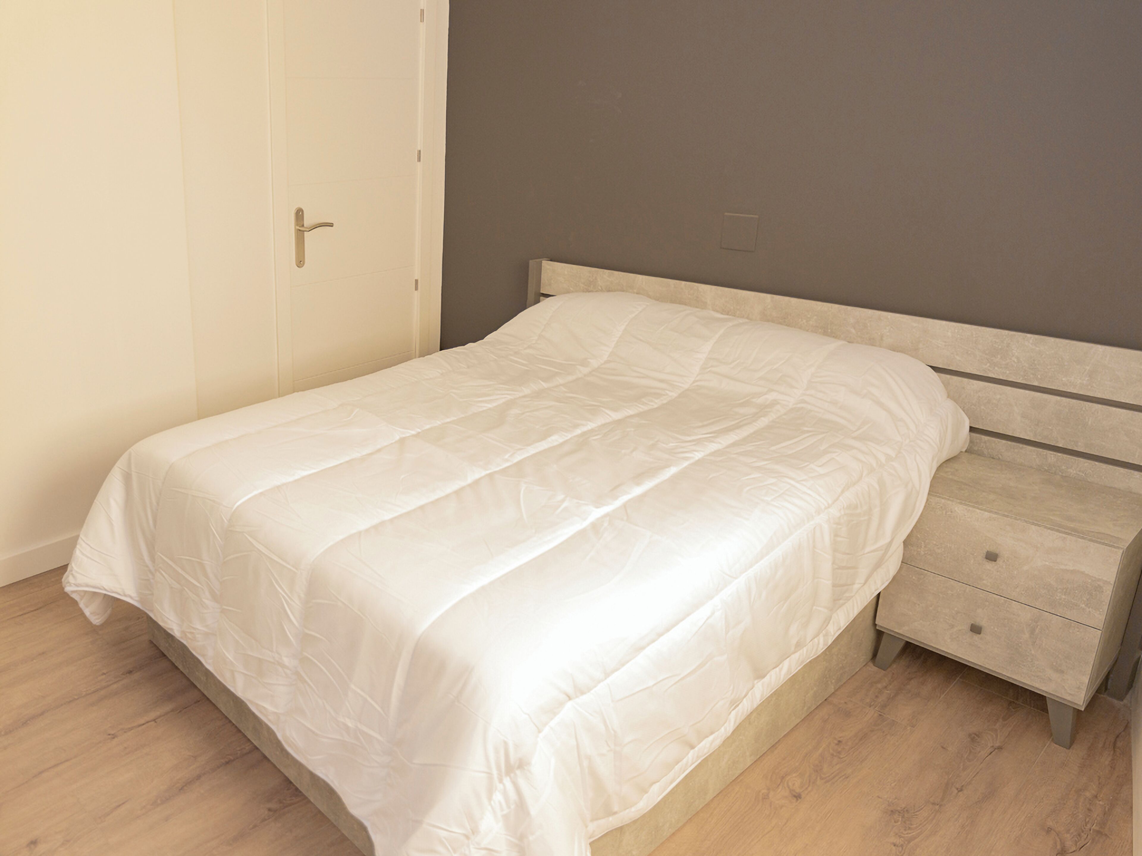 Una cama bien hecha con una colcha blanca y una mesita de noche de madera con cajones.