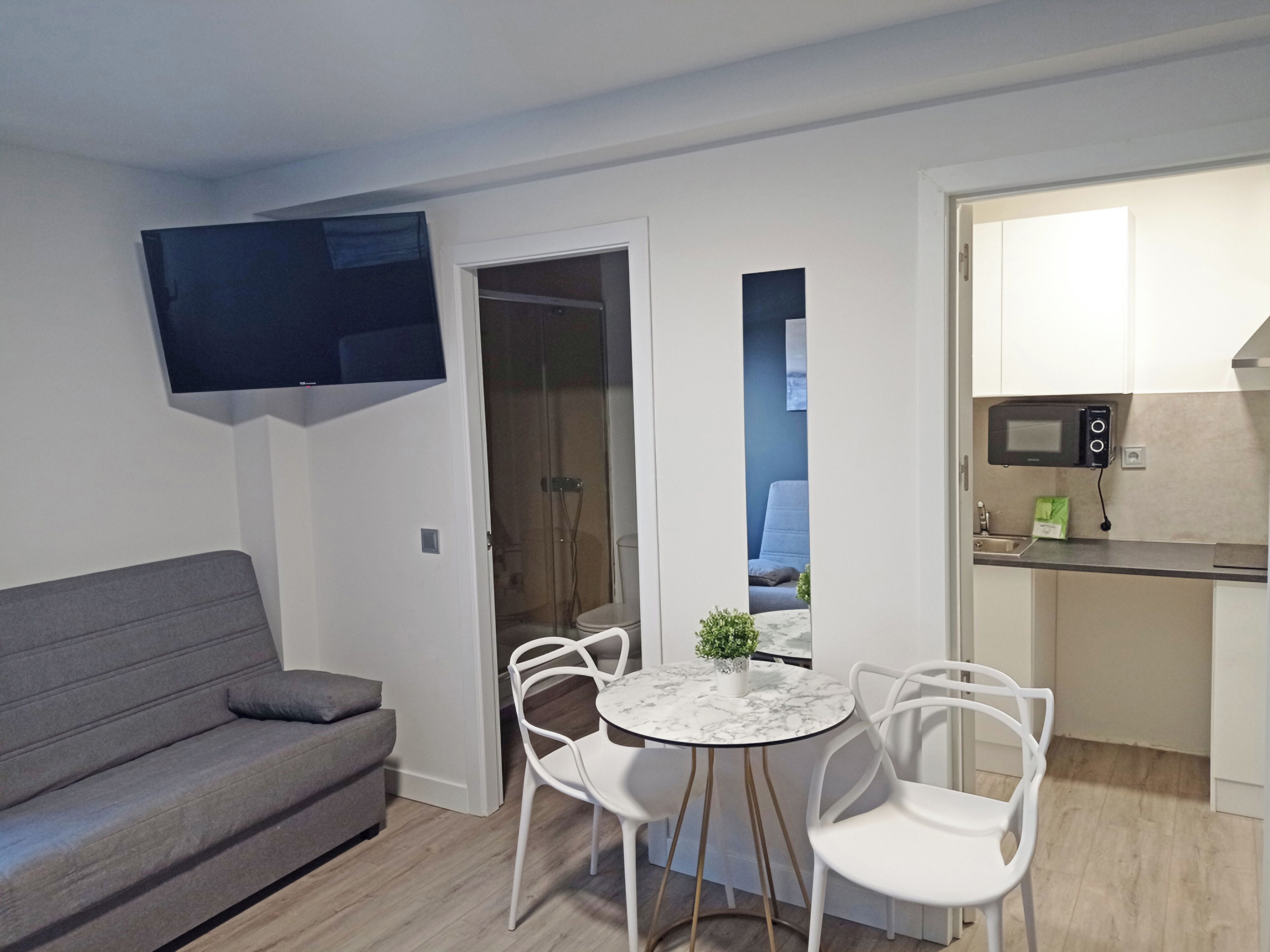 A compact living space with a grey sofa, a small round table with chairs, a TV mounted on the wall, and a kitchenette with a microwave.