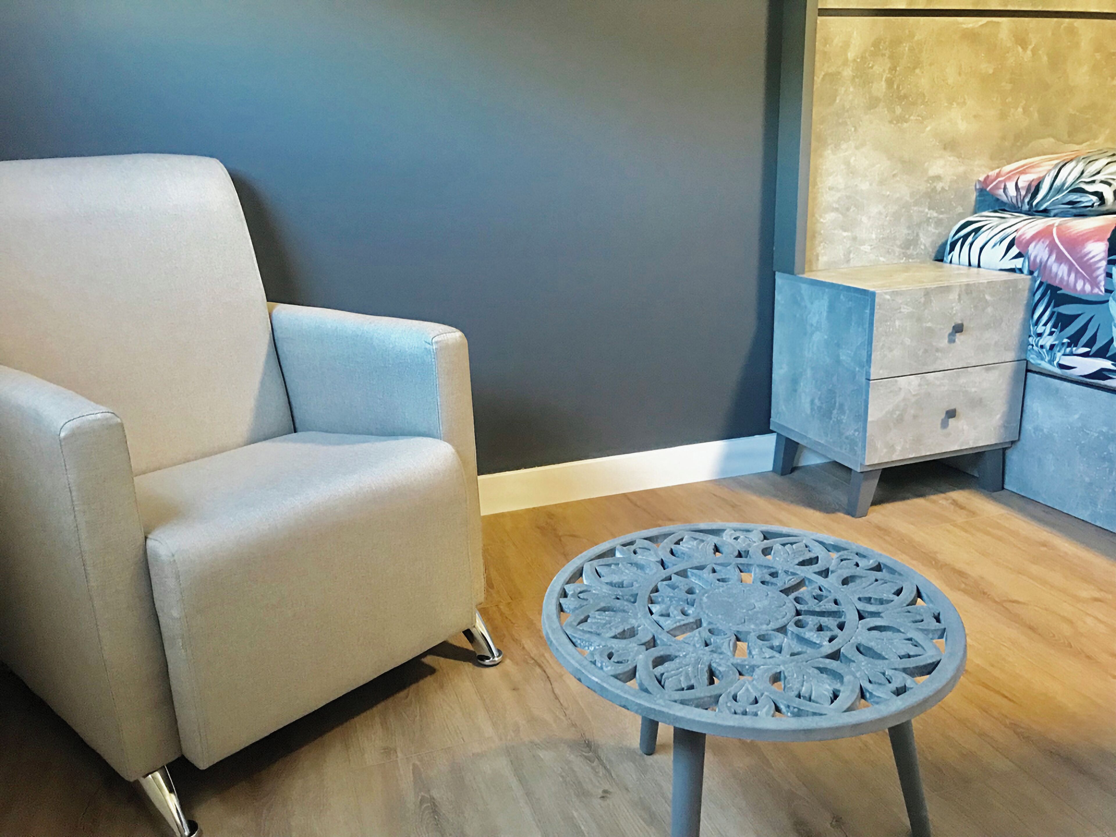 A room with a light blue armchair, a small round table with a decorative metal design, a nightstand with drawers, and a bed with a patterned blanket.