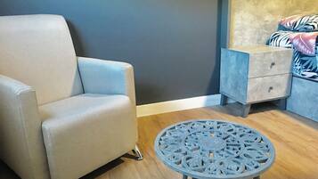 A room with a light blue armchair, a small round table with a decorative metal design, a nightstand with drawers, and a bed with a patterned blanket.