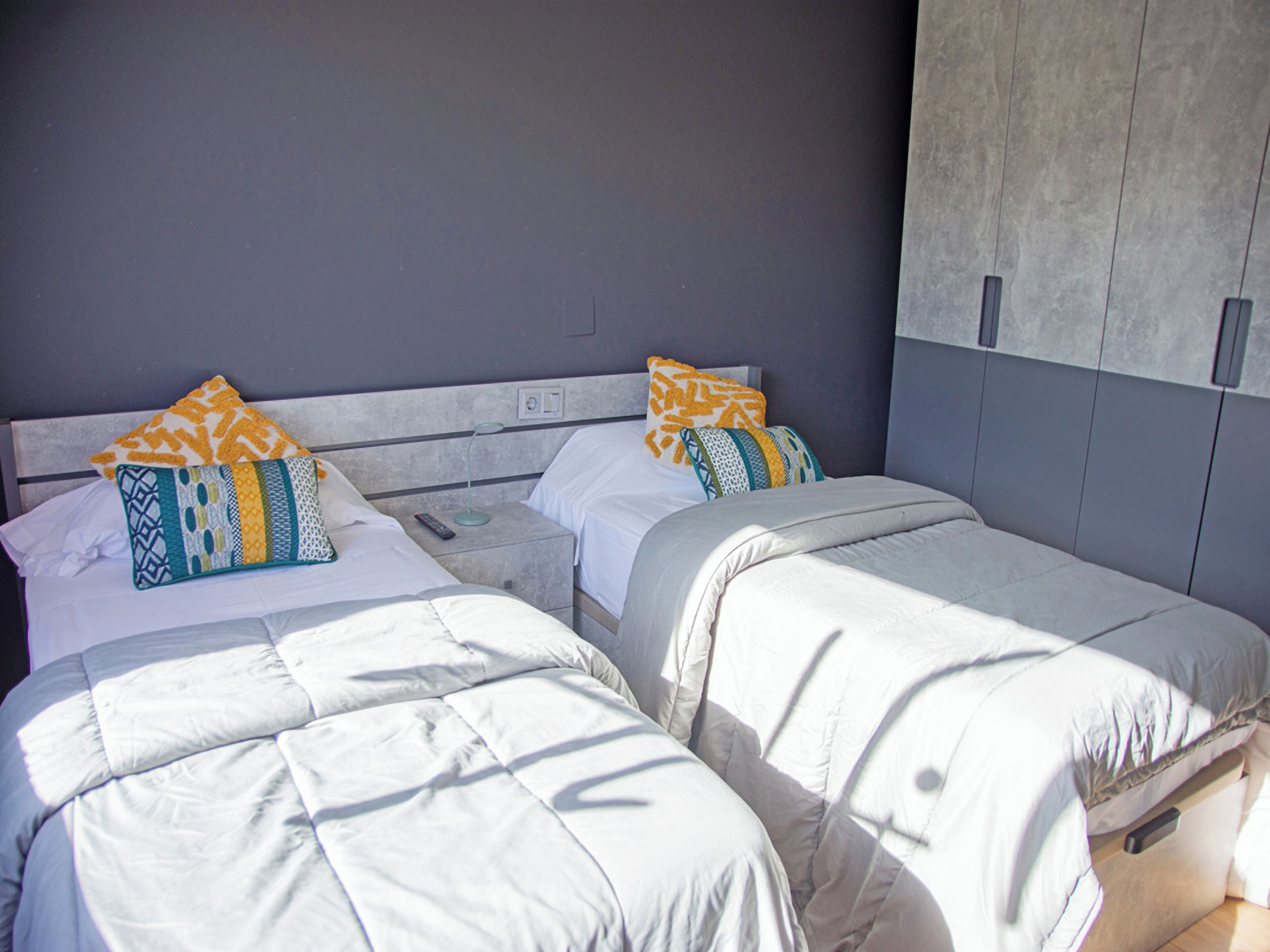Habitación de hotel con dos camas, cada una con ropa de cama blanca y almohadas estampadas. Hay una mesita de noche con una lámpara y un control remoto entre las camas. La habitación tiene una pared gris y un techo gris con una luminaria.