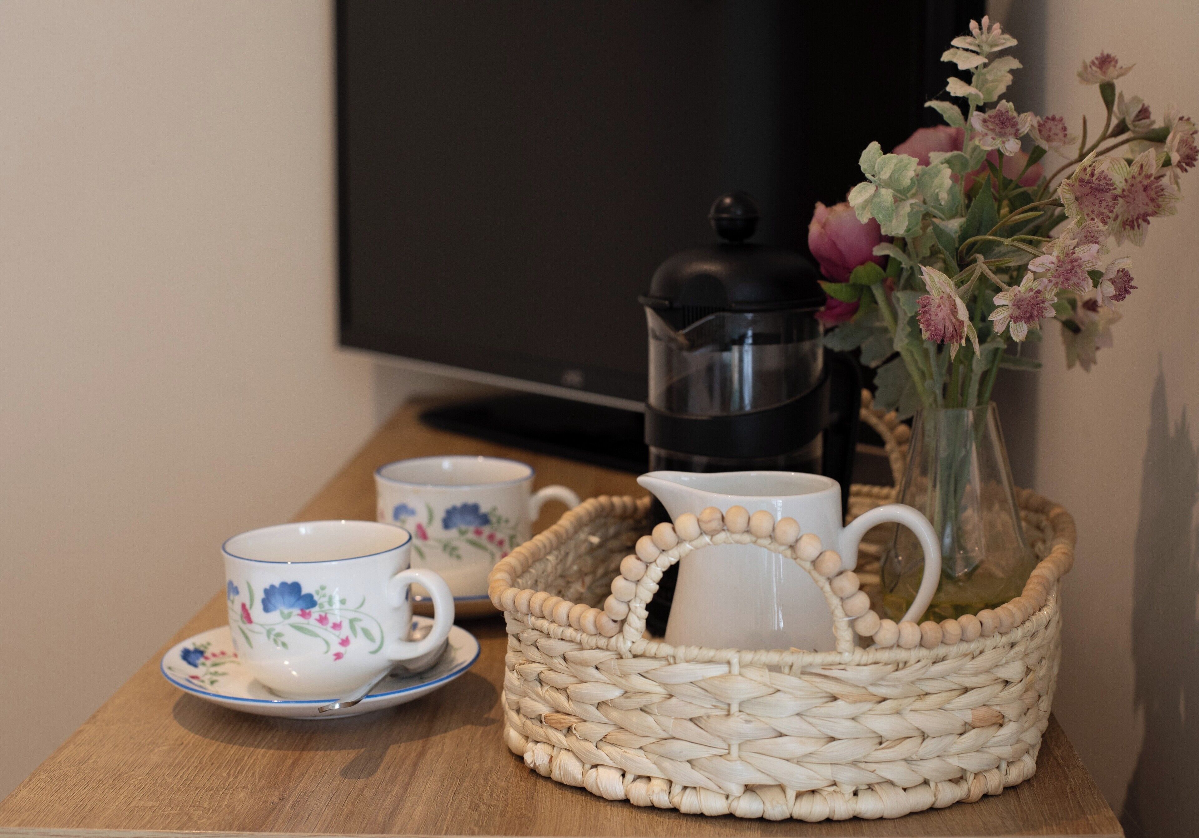 A table with a woven basket containing a white pitcher and a vase with flowers, and two floral-patterned cups with saucers.