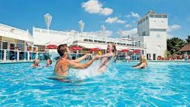Une piscine avec des personnes dans l’eau, un bâtiment à la façade blanche et un ciel bleu dégagé.