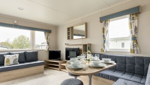 Une pièce moderne et bien éclairée, avec un canapé, un téléviseur, une table à manger dressée avec des assiettes et des verres, et de grandes fenêtres dotées de rideaux.