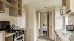Une cuisine étroite avec des meubles de rangement en bois, une cuisinière et un grille-pain. Un couloir mène à une chambre à coucher équipée d’un lit et d’une fenêtre.
