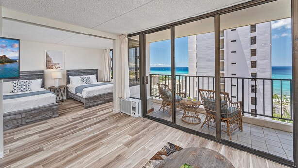 A balcony with a view of the ocean and a building. There are two beds with white bedding and a small table with a lamp.