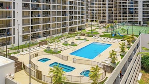 Um hotel com vários andares e área de piscina no terraço, incluindo uma piscina principal, piscinas menores e uma quadra de tênis.