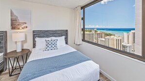 A hotel room with a bed, a nightstand, a lamp, a framed picture, and a large window with a city and ocean view.