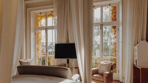 Un dormitorio con cama, una silla, televisión y ventanas amplias con cortinas.