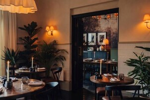 Interior de restaurante com mesas redondas, cadeiras, luminárias na parede e plantas em vasos.