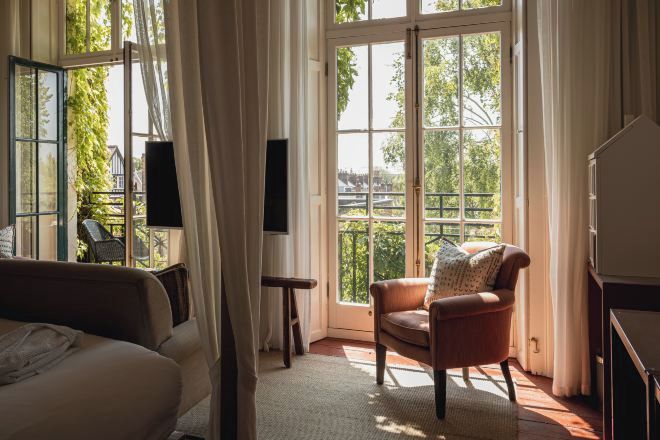 A room with a bed, a chair, a small table, and a large window with a view of greenery and buildings.
