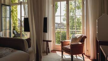 A room with a bed, a chair, a small table, and a large window with a view of greenery and buildings.