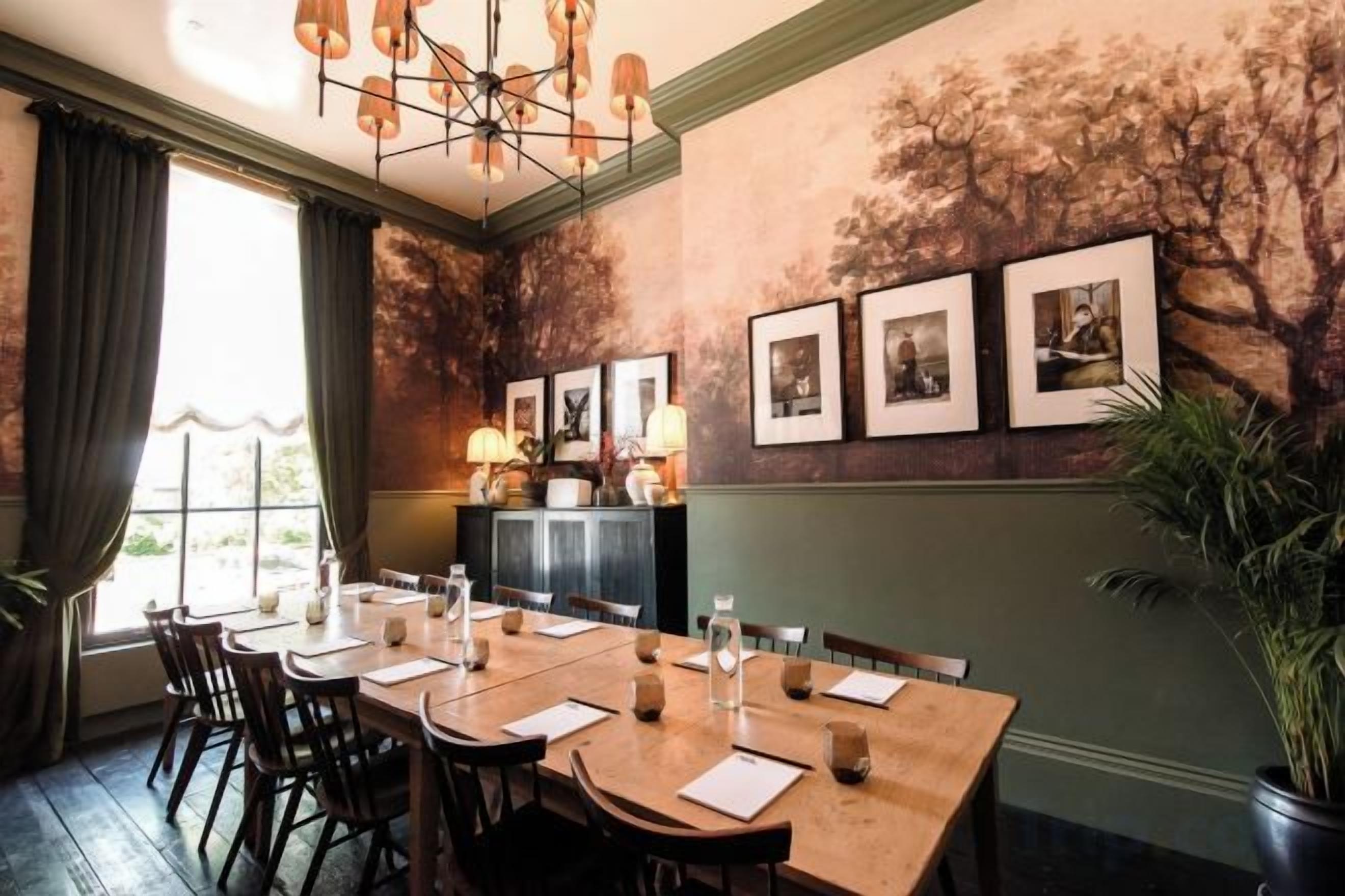 A dining area with a long table, chairs, and a chandelier. There are framed pictures on the wall and a plant in the corner.