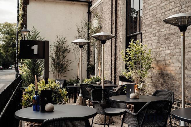 An outdoor cafe with round tables and chairs, potted plants, and a brick wall.