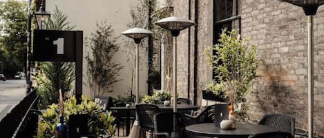 An outdoor cafe with round tables and chairs, potted plants, and a brick wall.