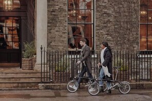 Deux personnes font du vélo en tandem devant un bâtiment doté de grandes fenêtres et d’une façade en briques.