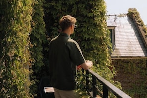 Une personne se tient sur un balcon, d’où l’on peut admirer un bâtiment en briques et un jardin.